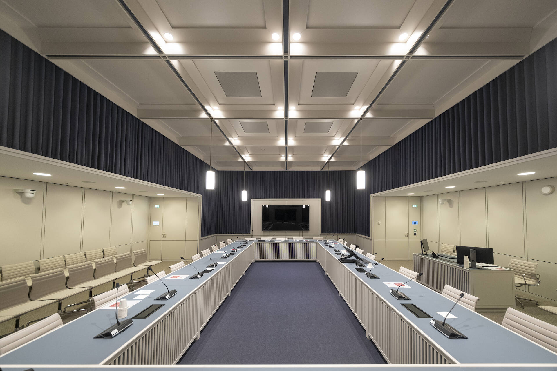 Op de afbeelding is commissiekamer 1 in het Eerste Kamergebouw te zien. De opstelling is een u-vormige tafel met stoelen eromheen.