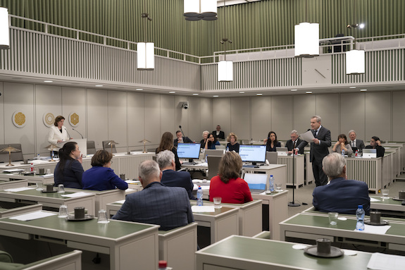 Foto van de plenaire vergaderzaal van de Eerste Kamer: mensen zitten in bankjes, een persoon staat bij de interruptiemicrofoon en een persoon staat achter het spreekgestoelte op de regeringstafel