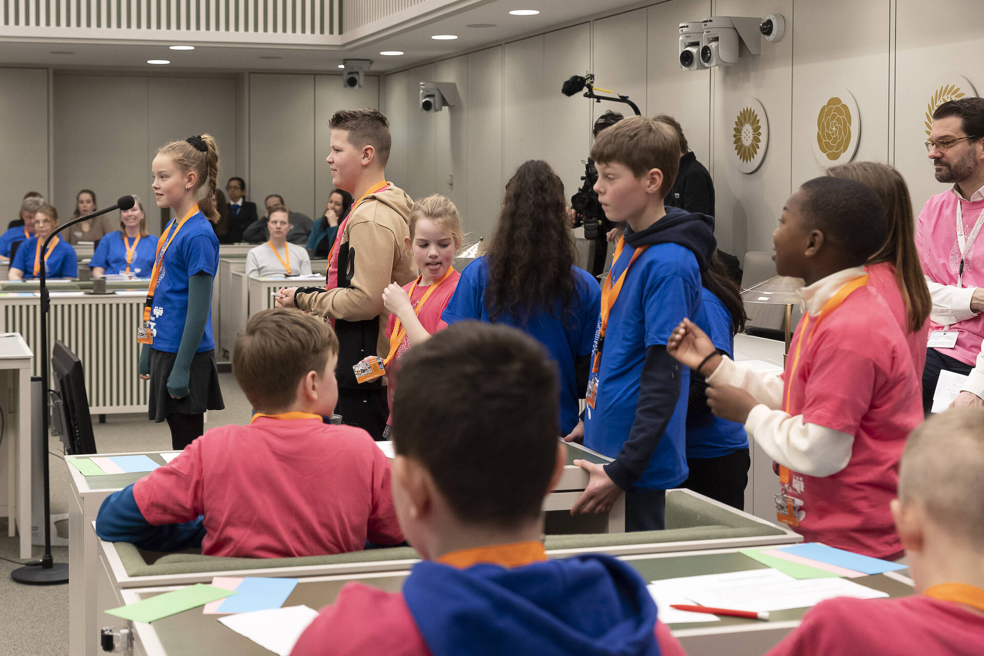 Een lange rij kinderen met roze en blauwe shirts staat achter elkaar bij een microfoon