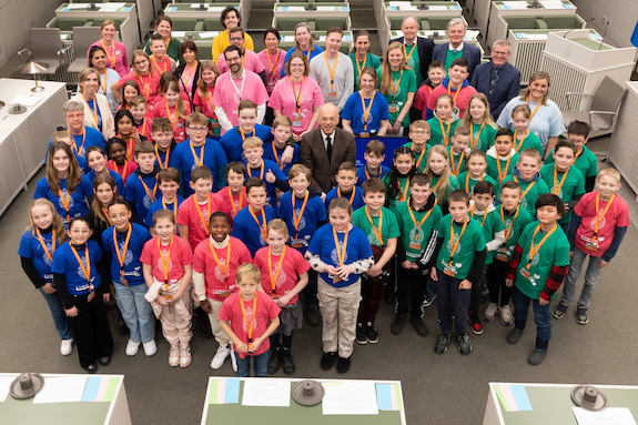 Foto van een grote groep kinderen en enkele volwassenen in de vergaderzaal van de Eerste Kamer. Ze dragen t-shirts in de kleuren roze, groen en blauw.