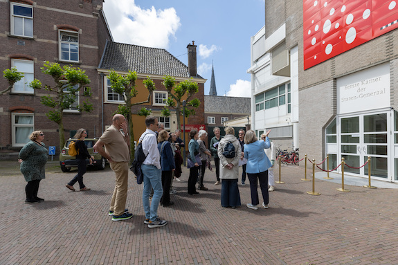 Foto van de gevel van de hoofdingang van de Eerste Kamer met daarvoor een groep mensen die uitleg krijgt over het kunstwerk aan de gevel